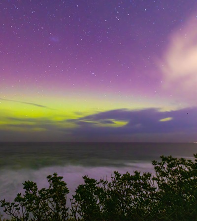 Aurora Australis or southern lights in Dunedin, New Zealand; beautiful purple, pink and green lights, with colourful clouds, fog ocean, night sky and mountains in the background